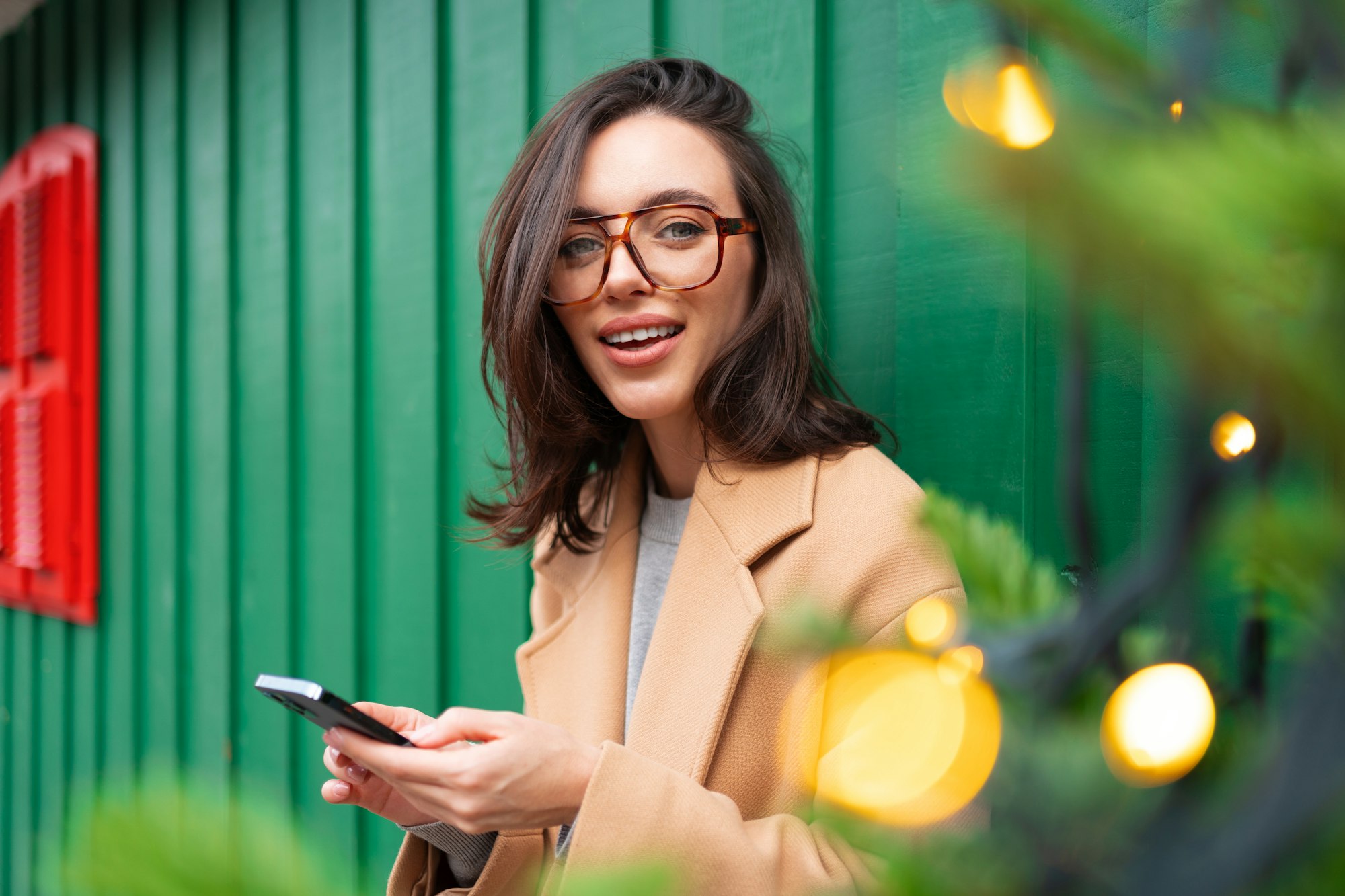 Close up smiling woman using smartphone, standing near Christmas tree